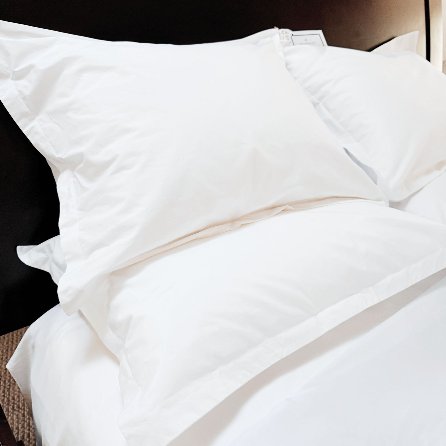 Kearsley, White 500 thread count Extra long staple cotton percale pillow shams with 3 cm flanges stacked on a dark brown bed. Kearsley Essentials Percale Pillowshams. Made in Italy.