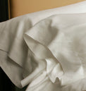 Kearsley Charlotte fine linens classic pillowcases.  Two shown in front of each other with a top sheet folded down and a tan leather head board in the back ground.