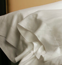 Kearsley Charlotte fine linens classic pillowcases.  Two shown in front of each other with a top sheet folded down and a tan leather head board in the back ground.